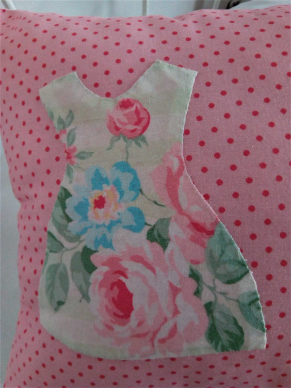 Petit coussin rectangulaire dimensions 26 X 45 cms Le fond du coussin est rose à petits pois Le coussin est agrémenté de 3 petites robes dans un tissu fleuri aux tons roses et jaunes, appliquées à petits points serrés La face arrière du coussin est blanche avec un petit coeur rose brodé main Matière du coussin 100 % coton Intérieur du coussin 100% polyester Conseil de lavage 30°